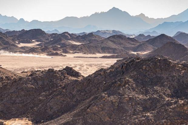 A panoramic view of a rugged desert landscape with distant mountains and a dusty trail.